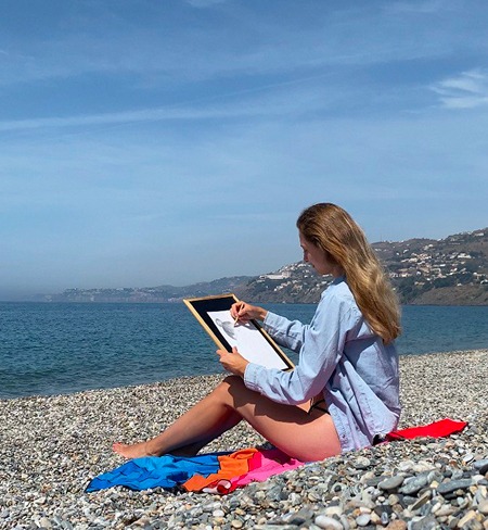 Artist Anneli is drawing a pencil portrait on a pebbled beach, enjoying the view of the sea. It's a sunny day, and she is wearing a blue shirt while sitting on a blanket, creating an atmosphere of relaxation and inspiration amidst nature.