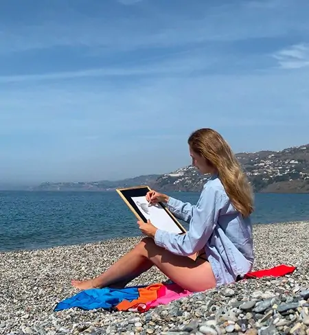 L’artiste Anneli de Anneli Studio dessine un portrait au crayon sur une plage de galets, profitant de la vue sur la mer. C’est une journée ensoleillée, elle porte une chemise bleue et est assise sur une couverture, créant une ambiance de détente et d’inspiration au cœur de la nature.