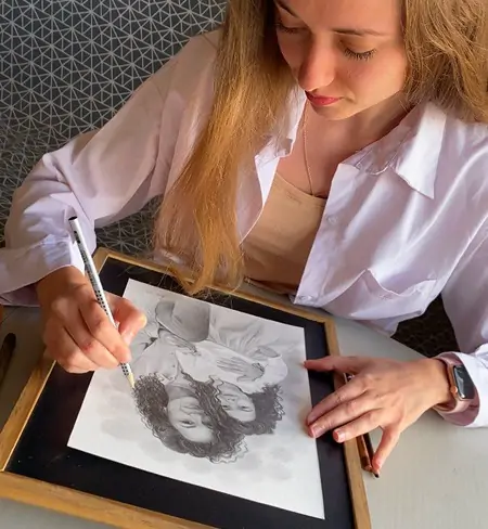L’artiste Anneli d’Anneli Studio dessine un portrait au crayon d’une mère et sa fille. Un gros plan révèle les lignes délicates et les détails fins de l’œuvre. Ses mains tiennent soigneusement le crayon, donnant vie à une image chaleureuse et pleine d’émotion.