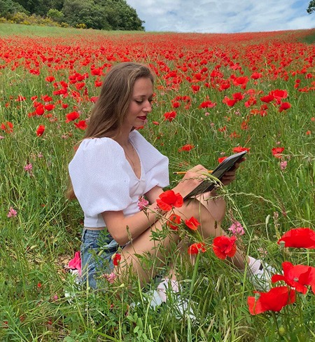 Artist Anneli is drawing a pencil portrait in a field of blooming poppies, wearing a white blouse with puffed sleeves. The vibrant red flowers surround her, creating a picturesque contrast with her outfit and adding a sense of spring and inspiration.