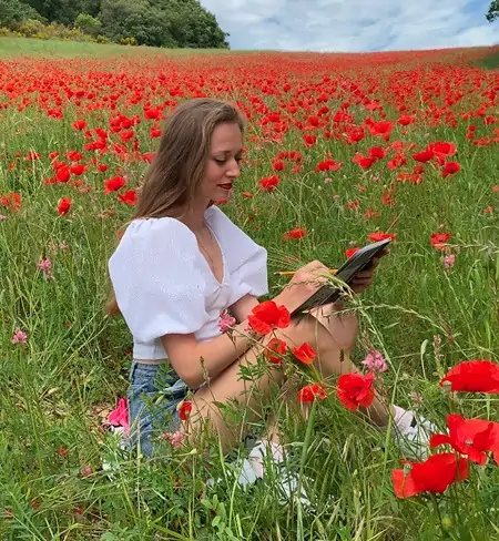 L’artiste Anneli de Studio Anneli dessine un portrait au crayon dans un champ de coquelicots en fleurs, vêtue d’un chemisier blanc à manches bouffantes. Les fleurs rouges éclatantes l’entourent, créant un contraste pittoresque avec sa tenue et apportant une sensation de printemps et d’inspiration.