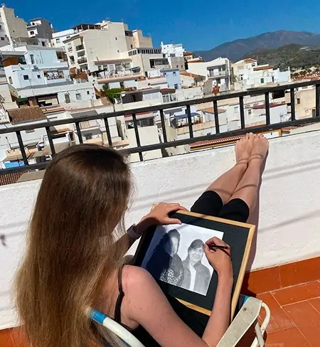 L’artiste Anneli Studio dessine un portrait au crayon sur une terrasse offrant une vue sur la ville andalouse de Salobreña, dans le sud de l’Espagne. Des maisons blanches dévalent la colline, baignées par une lumière chaude, créant une ambiance sereine et inspirante.