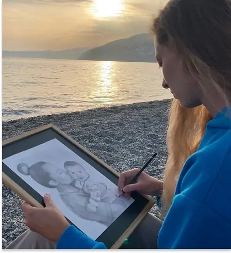 L’artiste Anneli de Anneli Studio dessine un portrait au crayon sur la plage au coucher du soleil. Derrière elle, un paysage pittoresque se déploie — mer calme, galets sous les pieds, et silhouettes de montagnes à l’horizon. Une atmosphère sereine et inspirante.