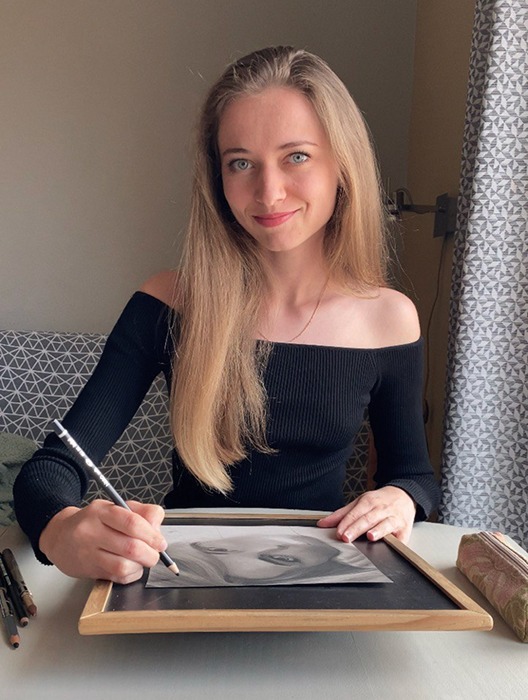 Artist Anneli is drawing a portrait at the table with pastels. She looks directly at the camera, smiling and creating a friendly atmosphere. She is wearing a black top with long sleeves and slightly off-shoulder, highlighting her style and confidence.