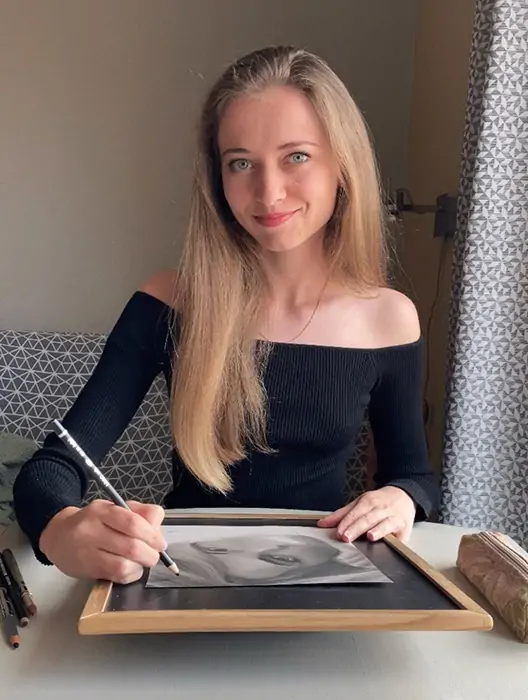 Artist Anneli is drawing a portrait at the table with pastels. She looks directly at the camera, smiling and creating a friendly atmosphere. She is wearing a black top with long sleeves and slightly off-shoulder, highlighting her style and confidence.