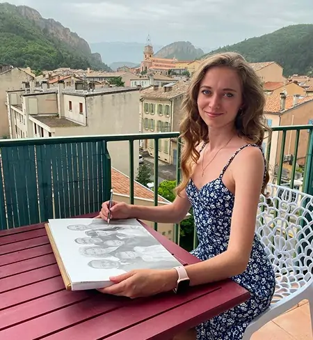 L’artiste Anneli dessine un grand portrait de famille au crayon sur un balcon ouvert, avec vue sur les montagnes de la ville de Digne-les-Bains en France. Elle est magnifique dans une robe, coiffée avec élégance, souriant à la caméra, créant une ambiance chaleureuse et conviviale.