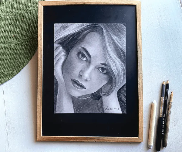 Le portrait d’Emma Stone, réalisé au fusain sur papier gris teinté, est exposé sur la table. À proximité, des crayons sont soigneusement rangés, prêts à être utilisés. Cette mise en scène crée une ambiance élégante et artistique, mettant en valeur les détails et la texture de l’œuvre.