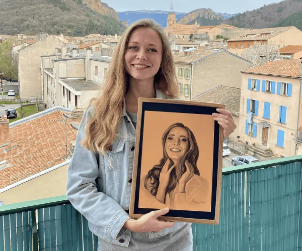 L’artiste Anneli tient son autoportrait sur le balcon avec une vue imprenable sur les montagnes. L’autoportrait, réalisé au pastel sur papier ton pêche, la montre en train de sourire, reflétant la même émotion joyeuse que dans la réalité. Cela crée une harmonie entre l’art et la vie.