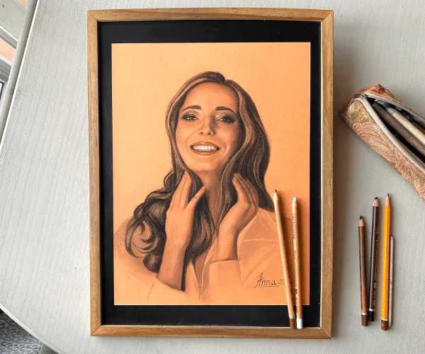 Self-portrait of Anneli, created in pastel on toned peach paper, depicts her laughing. Warm brown tones create a cozy atmosphere, highlighting her expression. Next to the portrait, pastel pencils used for the artwork are neatly arranged.