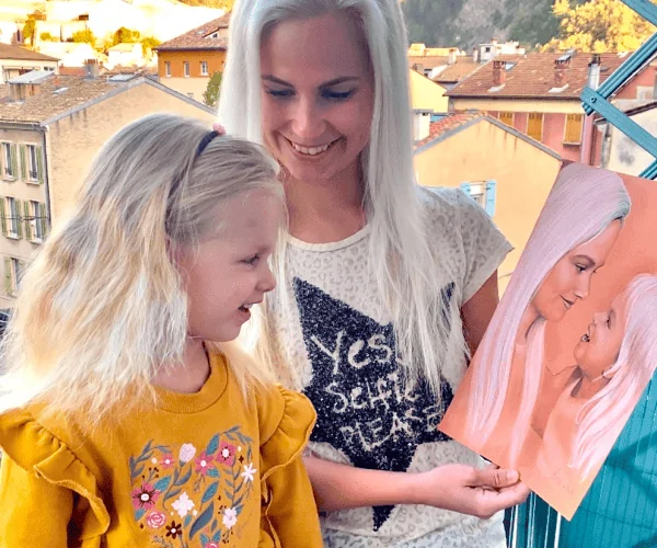 A mother and daughter joyfully hold a portrait of themselves. The portrait, created in pastel on toned peach paper, depicts the young mother and her three-year-old daughter looking at each other with sincere smiles. Both are joyful, with white hair that unites them. The portrait is done in several colors, creating a vibrant and warm impression.